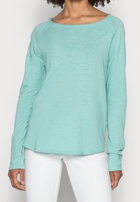 Langärmliges Shirt in sanftem Türkis, aus weichem, strukturiertem Stoff. Mit weitem Ausschnitt und abgerundetem Saum, kombiniert mit weißen Jeans.