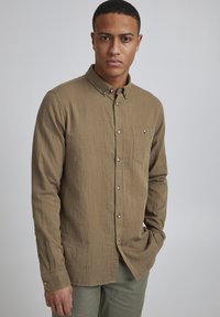 Chemise marron à manches longues en tissu texturé, avec une poche poitrine unique et des boutons argentés. Design sobre, style minimaliste.