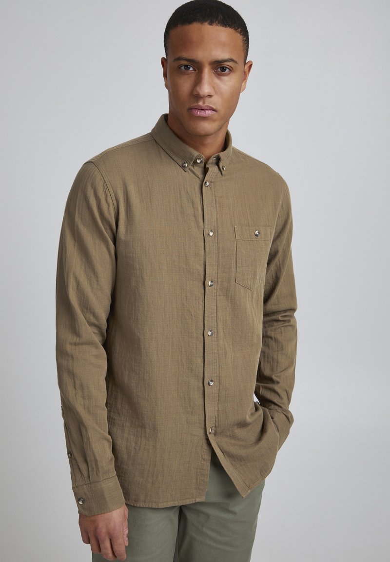 Chemise marron à manches longues en tissu texturé, avec une poche poitrine unique et des boutons argentés. Design sobre, style minimaliste.