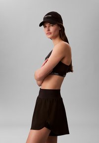 Femeie purtând un sutien sport negru Calvin Klein, fustă neagră și șapcă neagră Calvin Klein, stând cu brațele încrucișate pe un fundal gri simplu.