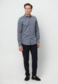 Camicia button-up in chambray blu con colletto, maniche lunghe e bottoni scuri, indossata con pantaloni scuri e scarpe in suede marroni.