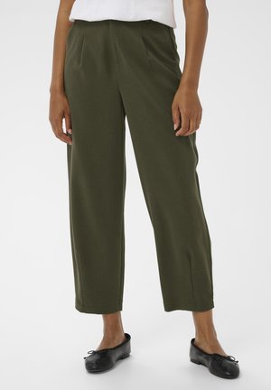 Frau trägt olivgrüne, hoch taillierte Hose mit weitem Bein, weißes Oberteil und schwarze Ballerinas, steht vor weißem Hintergrund.