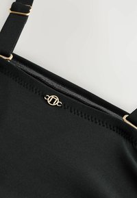 Juodas audinio dirželis su reguliuojamu auksinės spalvos sagtimi ir mažu apskritu aukso logotipo detaliu, esančiu netoli siūlės krašto.