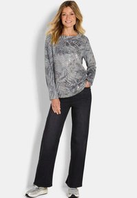Vrouw in een grijs gemarmerd langshirt, zwarte wijde jeans en grijze sneakers, lachend met één hand in haar zak.