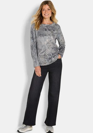 Frau trägt ein graues, gemustertes Langarmshirt, schwarze weit geschnittene Jeans und graue Turnschuhe, lächelt und hat eine Hand in der Tasche.