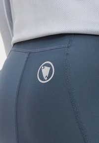 Κοντινό πλάνο μπλε αθλητικών κολάν με ενισχυμένες ραφές και κυκλικό ανακλαστικό λογότυπο στο πάνω πίσω μέρος του μηρού.