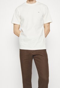 Bílá bavlněná trička s krátkým rukávem a kulatým výstřihem, s malým černým logem na levé horní části hrudi. Spárováno s hnědými kalhotami.