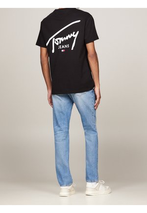 Person iført sort "Tommy Jeans" T-shirt og lyseblå jeans, stående med ryggen mod en neutral baggrund.