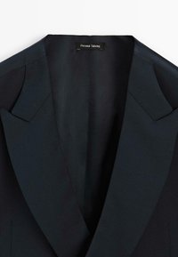 Dunkelblauer Blazer mit Reverskragen, taillierter Passform und glatter Textur. Das Innenetikett trägt die Aufschrift "Maßgeschneiderte Anfertigung".