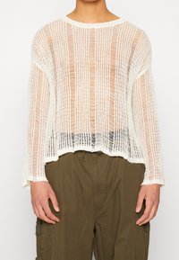 Person som bär en vit, löst stickad långärmad tröja över en bar överkropp, kombinerad med höga olivgröna cargobyxor.