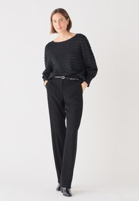 Schwarzer, strukturierter Pullover mit weitem Ausschnitt, kombiniert mit hochtaillierten schwarzen Hosen. Ein subtiler Gürtel und Ankle Boots runden das Outfit ab.