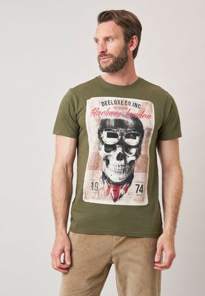 Homme avec barbe portant un t-shirt vert olive avec un grand motif de crâne, portant des lunettes et une écharpe, regardant sur le côté sur un fond uni.