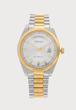 Montre-bracelet automatique bicolore argent et or Versace avec cadran rond blanc, index dorés, affichage du jour et de la date, et bracelet à maillons métalliques.