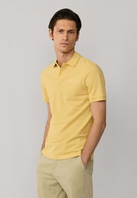 Homme portant un polo à manches courtes jaune et un pantalon beige, debout les mains dans les poches devant un fond gris clair uni.