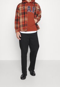 Camisa de cuadros en naranja, marrón y negro, superpuesta a una sudadera marrón con letras blancas. Unos pantalones negros y botas negras completan el atuendo.