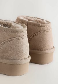 Beige suede ankelstøvler med blødt pelsfor og holdbare gummisåler, vist fra baghånd på en lys baggrund.