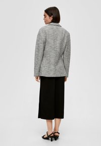 Frau von hinten, die ein graues, strukturiertes Blazerjacke, schwarze, weit geschnittene, knöchellange Hose und schwarze Absatzsandalen trägt, vor einem schlichten Hintergrund.
