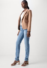Blazer marrone aderente su una maglietta bianca, abbinato a jeans azzurri chiari e sandali beige con lacci. Tessuto morbido, design sartoriale e bottoni notevoli.