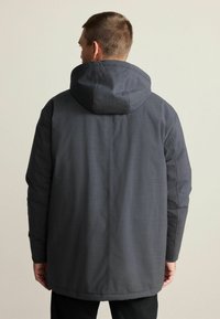 Ein Mann trägt eine dunkelgraue Kapuzenjacke, gesehen von hinten vor einem schlichten beigen Hintergrund.