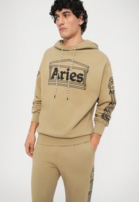 Beżowa bluza z kapturem z grafiką "Aries" w kolorze czarnym, z regulowanym kapturem i detalami na rękawach; w komplecie ze zgodnymi beżowymi spodniami dresowymi.