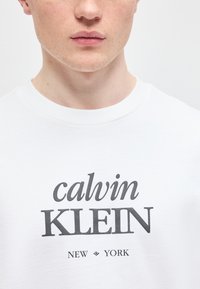 Νεαρός/Νεαρή φοράει λευκό μπλουζάκι με την επιγραφή "calvin KLEIN NEW YORK" τυπωμένη με μαύρα γράμματα στο στήθος.