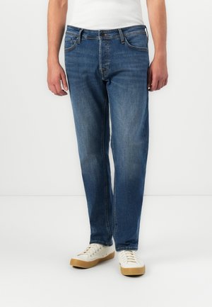 Man in blauwe recht gesneden jeans en witte sneakers, staand tegen een effen witte achtergrond.