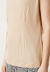 Beige mouwloze top van een zachte, gestructureerde stof met subtiele glans, zijplooien en een afgeronde zoom, gecombineerd met zwart-witte gestreepte broek.
