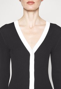 Cardigan nero a coste con scollo a V profondo, caratterizzato da un bordo bianco e chiusura a bottoni sul davanti. Texture morbida e design sagomato.