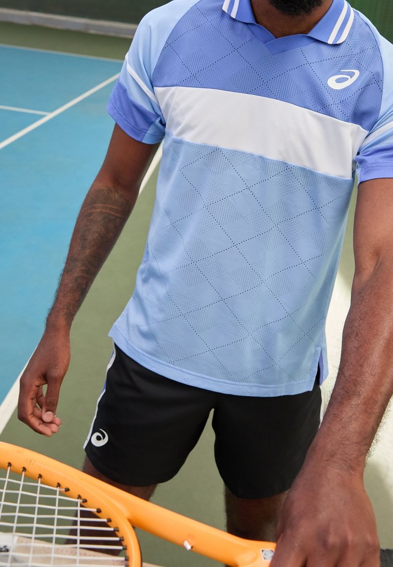 Jasnoniebieska koszulka sportowa z białym paskiem, z diamentowym wzorem i dzianinową teksturą. W połączeniu z czarnymi szortami i pomarańczową rakietą tenisową.