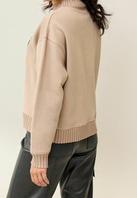 Sweat-shirt beige avec un col montant côtelé, des poignets et un ourlet côtelés. Le tissu semble doux, avec une coupe décontractée. Porté avec un pantalon noir en similicuir.