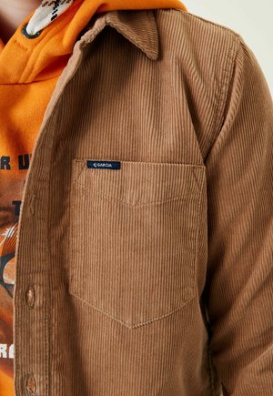 Camisa de pana marrón con un bolsillo en el pecho y una etiqueta "Garcia". Debajo, se ve parcialmente una sudadera con capucha naranja con un estampado gráfico.