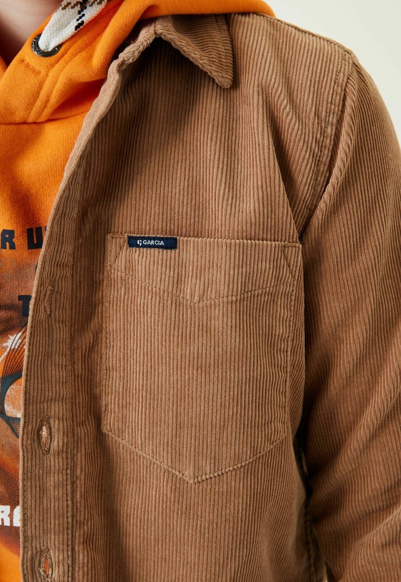 Chemise en velours côtelé marron avec une poche poitrine et une étiquette "Garcia". En dessous, un sweat à capuche orange avec un imprimé graphique est partiellement visible.