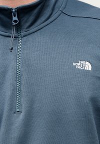 Φλις πουλόβερ σε σκούρο μπλε χρώμα με ψηλό γιακά και φερμουάρ τετάρτου μήκους, διακοσμημένο με λευκό λογότυπο στο στήθος και υφή υφάσματος με ανάγλυφη επιφάνεια.