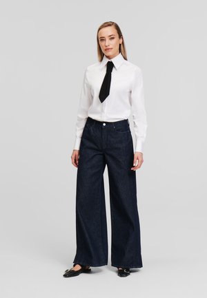 Witte overhemden met een zwarte stropdas, gecombineerd met donkerblauwe jeans met een wijde pijp. Zwarte platte schoenen maken de outfit compleet.