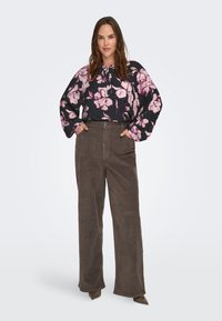 Czarna bluzka w kwiaty z różowymi kwiatami, długie rękawy; w połączeniu z brązowymi, szerokimi spodniami z sztruksu o wysokim stanie. Całość dopełniają buty z wydłużonym noskiem.