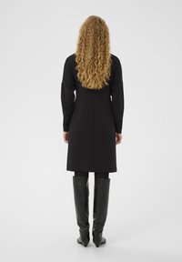 Czarna, długa do kolan sukienka z długimi rękawami, wykonana z gładkiego materiału, zestawiona z wysokimi, dopasowanymi czarnymi skórzanymi botkami. Kręcone blond włosy.