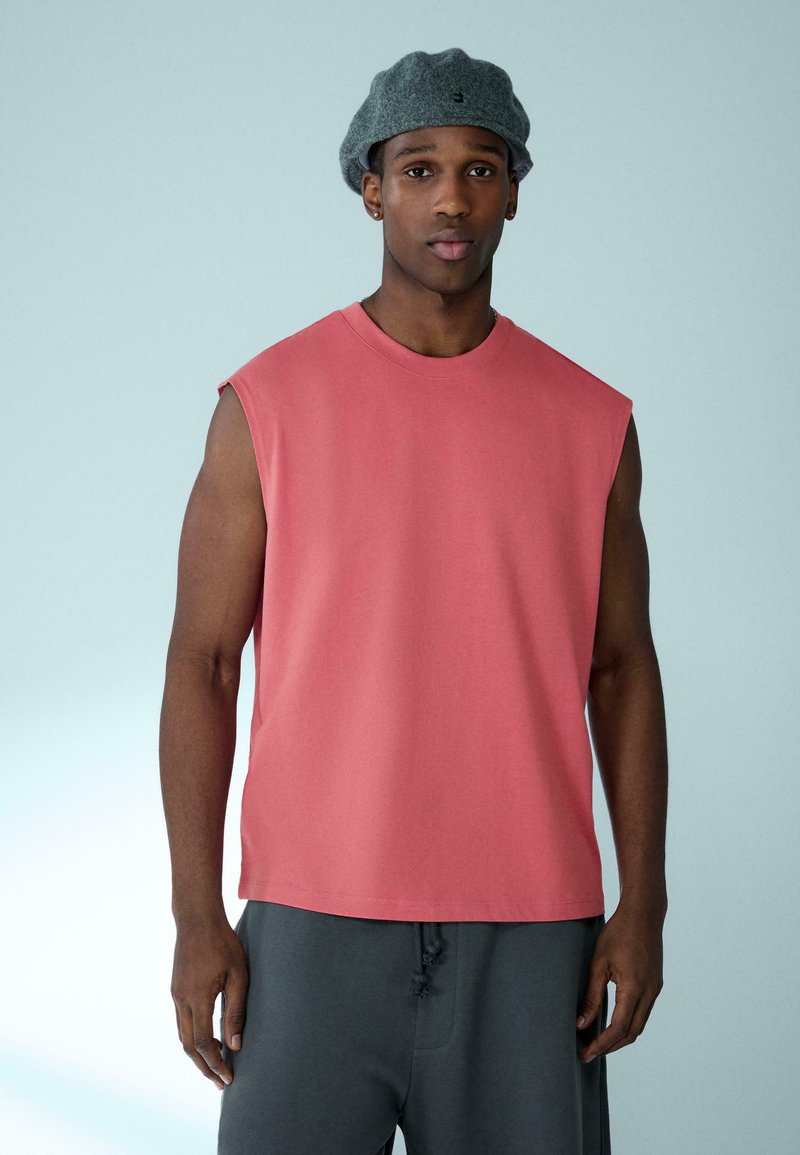 T-shirt sans manches en coton couleur corail avec un col rond. Porté avec un short gris foncé. Texture lisse et coupe décontractée.