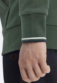 Felpa verde scuro con polsini a coste caratterizzati da una striscia bianca e verde scuro. Mano chiusa a pugno, che mostra la texture e i dettagli della pelle.