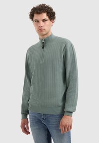 Lichtgroene gestructureerde trui met een hoge hals en een halve rits. Lange mouwen en ribgebreide boorden. Geweven patroon over de hele trui. Gecombineerd met spijkerbroek.