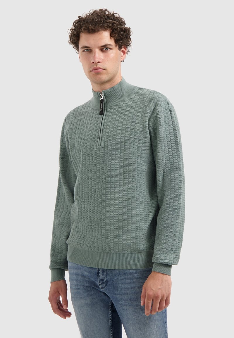 Lichtgroene gestructureerde trui met een hoge hals en een halve rits. Lange mouwen en ribgebreide boorden. Geweven patroon over de hele trui. Gecombineerd met spijkerbroek.