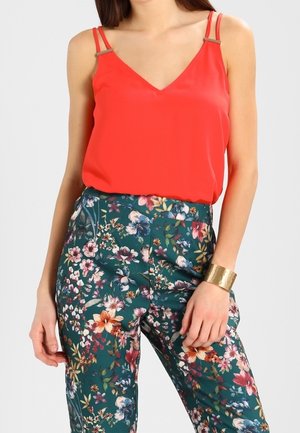 Vrouw die een rood mouwloos topje met dubbele bandjes draagt en een groene hoge taille broek met bloemenprint, afgewerkt met een brede gouden armband.