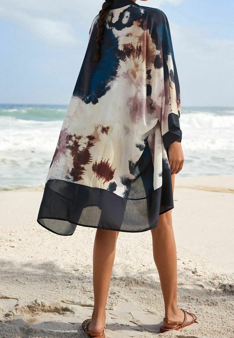 Personne portant un couvre-chef ample multicolore tie-dye, debout pieds nus sur une plage de sable avec les vagues de l’océan en arrière-plan.