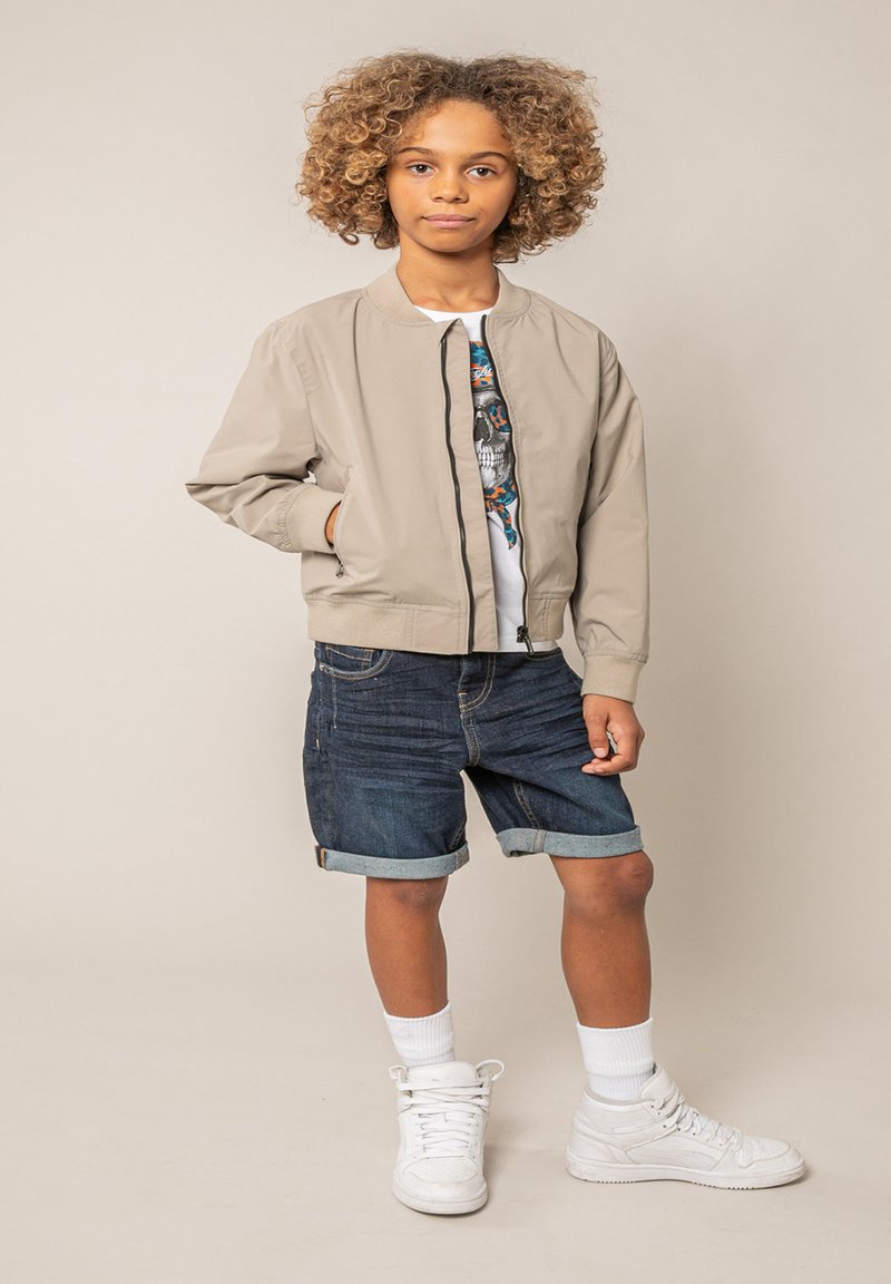 Enfant aux cheveux bouclés portant une veste aviateur beige, un T-shirt graphique, un short en denim retroussé, des chaussettes blanches et des baskets blanches, debout devant un fond uni.