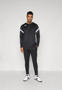 Fekete sportos hosszú ujjú felső fehér vállcsíkkal és hozzáillő nadrág, fehér sportcipővel, amelyen fekete részletek vannak.