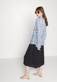 Blusa floral azul clara com mangas franzidas, combinada com uma saia midi preta e sandálias brancas. Os óculos de sol são oversized com uma armação rosa.
