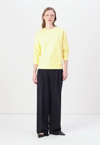 Geel katoenen sweatshirt met lange mouwen en ribbelmanchetten, gecombineerd met een hooggewaisted zwarte wijde broek en puntige zwarte schoenen.