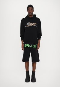 Haut à capuche noir avec un motif léopard ; short noir avec un accent graphique vert ; modèle portant des bottes noires.