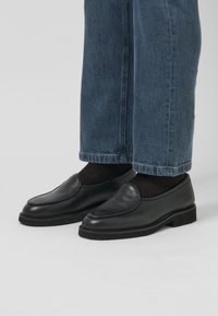 Černé kožené mokasíny s texturovaným svrškem, kulatou špičkou a nízkou gumovou podrážkou, spárované s volnými modrými denimovými džínami.