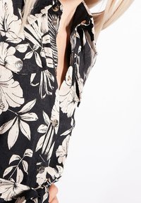 Czarna koszula z dużym białym nadrukiem kwiatowym, widocznymi guzikami i częściowo rozpiętym kołnierzykiem, noszona przez osobę z blond włosami.