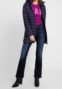 Marinblå puffjacka med horisontellt quiltat mönster, bärs över en fuchsia skjorta med vita bokstäver, kombinerat med mörka utsvängda jeans och svarta klackar.